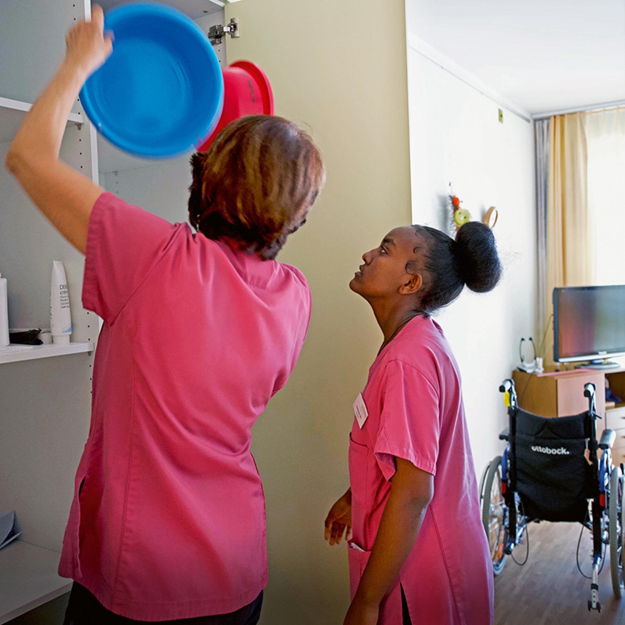 La foto mostra una scena in una camera di una casa per anziani: un’assistente di cura mostra a una giovane donna cosa c’è da fare.