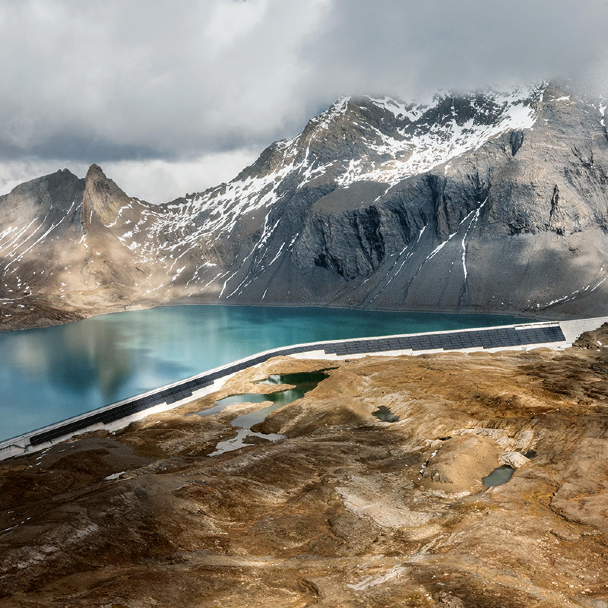 La foto mostra un paesaggio di montagna con un lago artificiale sulla cui diga sono istallati pannelli solari.